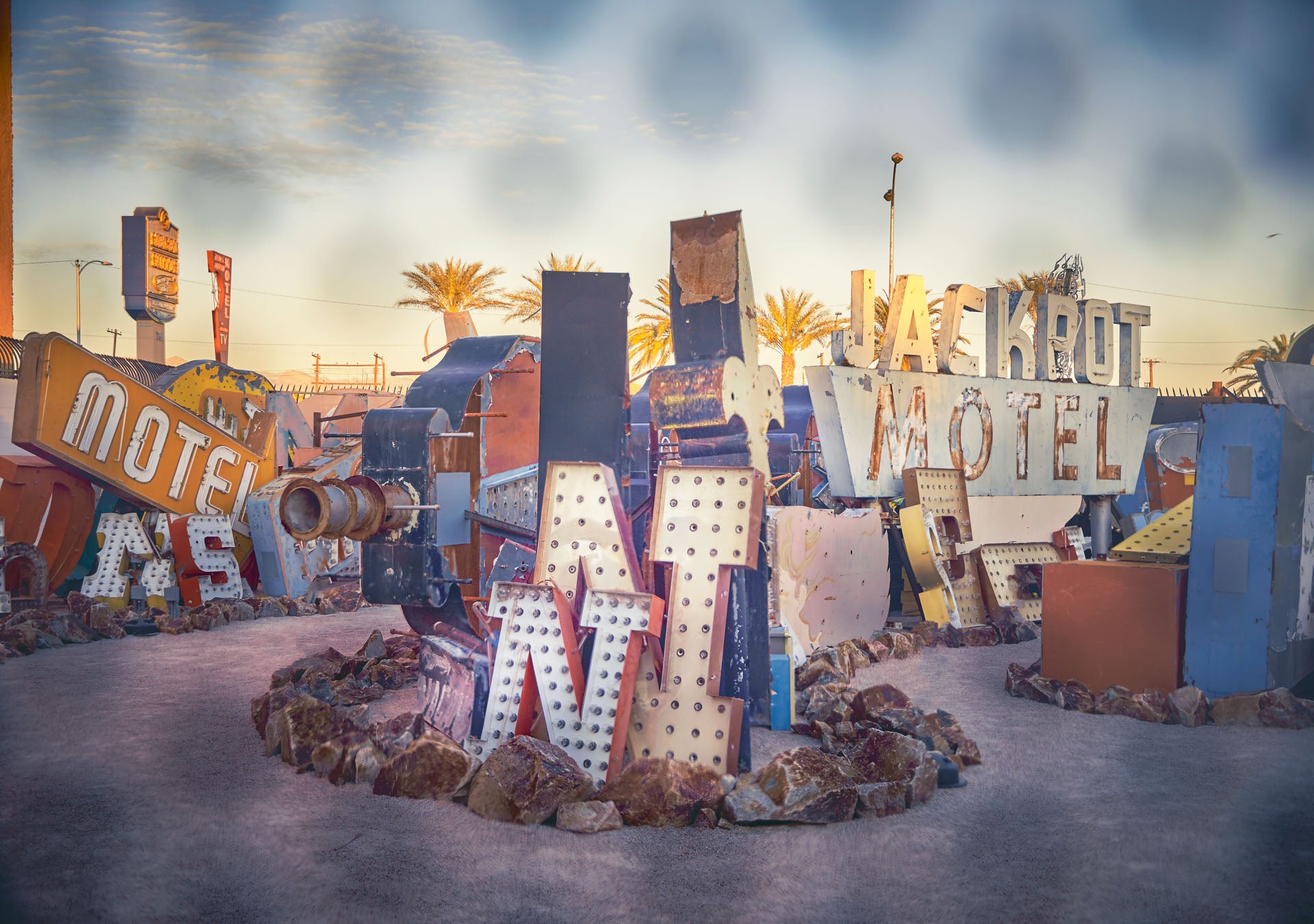 Vintage Las Vegas neon boneyard at golden hour with rusted “JACKPOT MOTEL” sign, scattered bulb-studded letters, palms, and a curving gravel path.