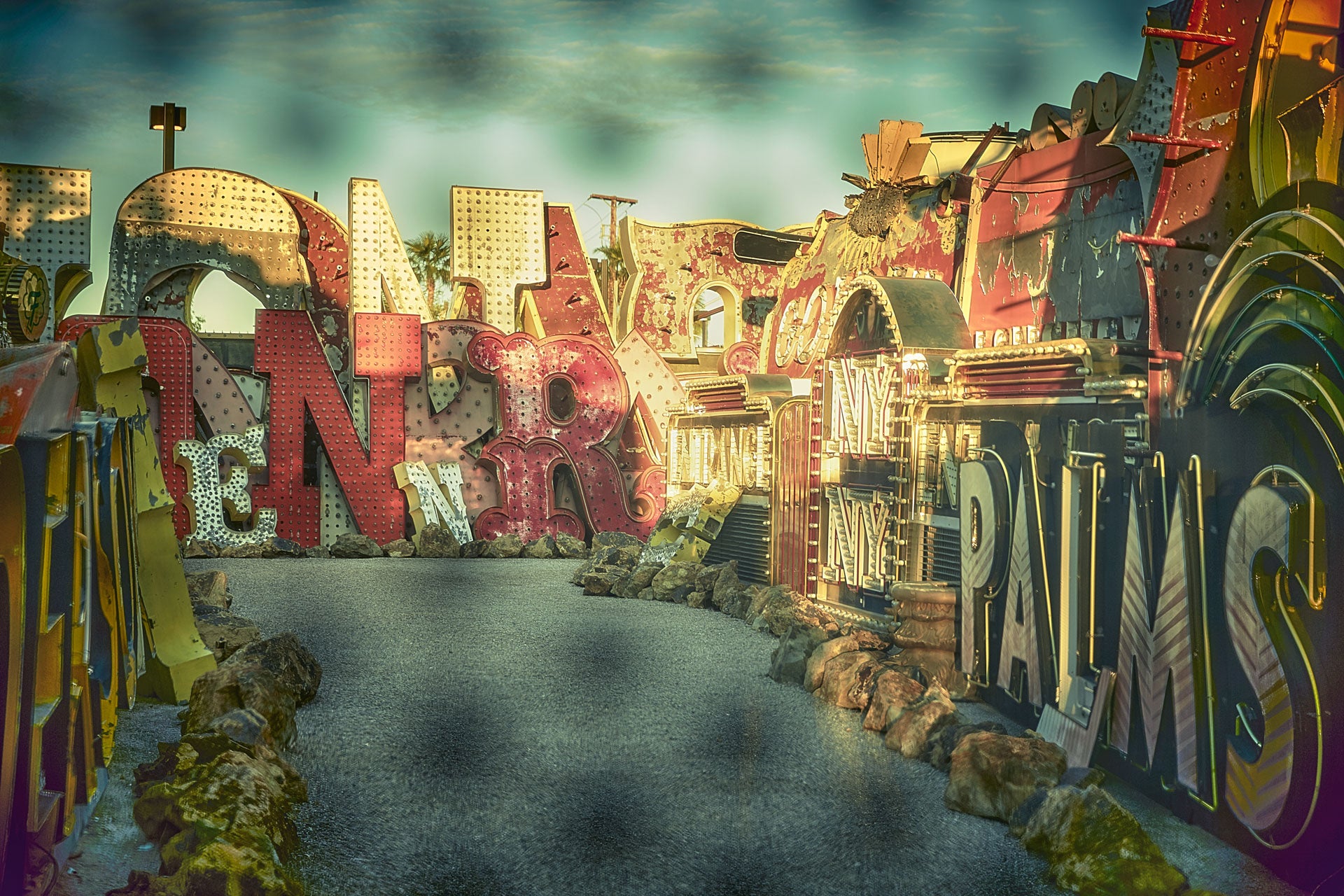 Sunlit alley of vintage Las Vegas casino signs—layered neon letters, riveted metal, and weathered paint (“NYNY,” “PALMS”) arranged along a curving path.