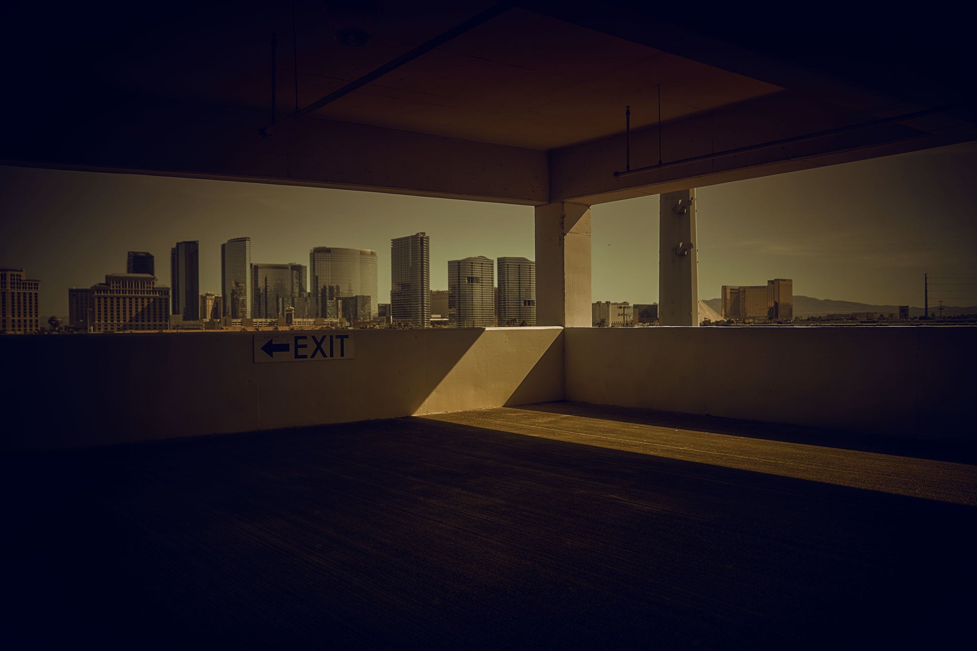 Framed view from an upper parking garage toward the Las Vegas skyline; “EXIT” sign on a concrete wall and a long diagonal shadow across the empty deck in warm desert light.
