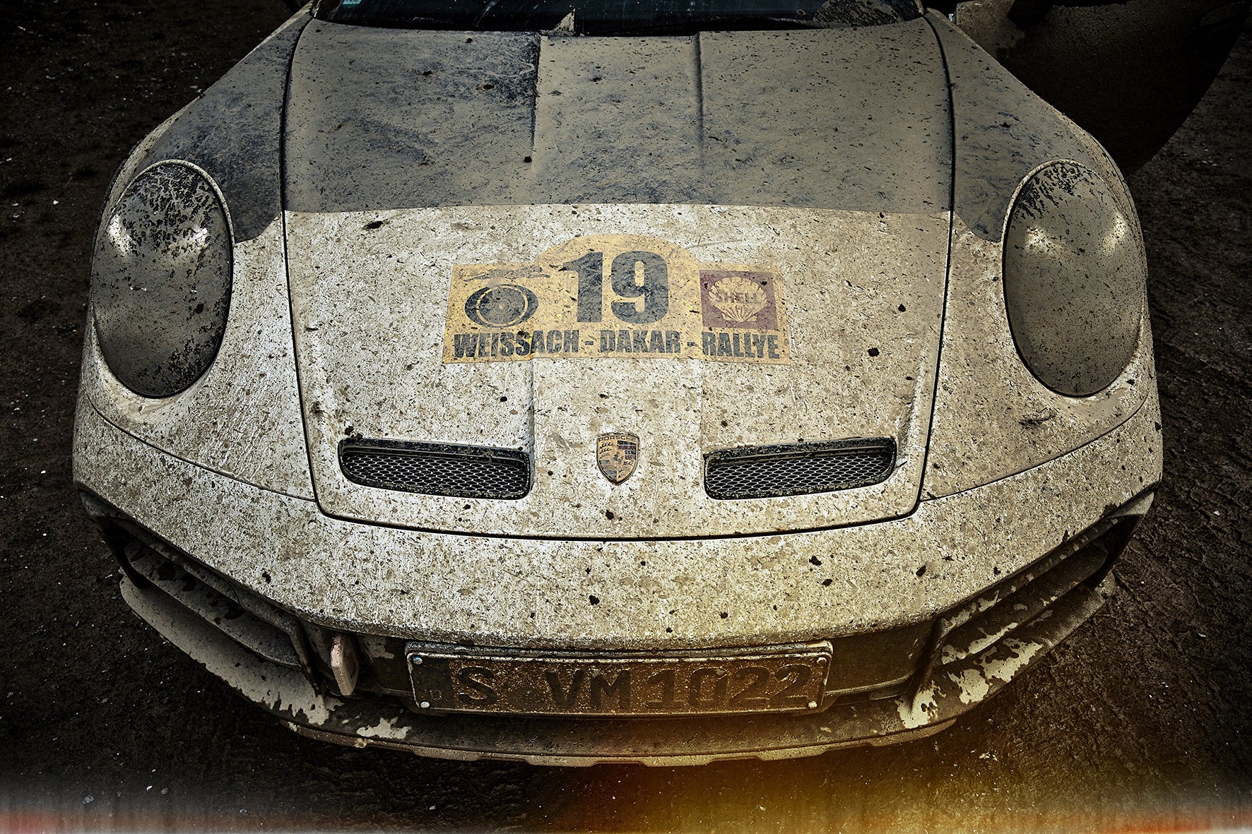 Mud-caked front of a Porsche 911 Dakar with “19 – Weissach · Dakar · Rallye” hood sticker; spattered headlights, badge, and grilles in gritty detail.