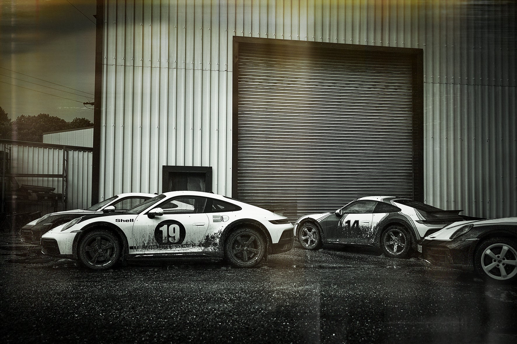 Fine art photo by Frank Kayser – Porsche Dakar cars lined up after a day in mud, industrial setting in background.