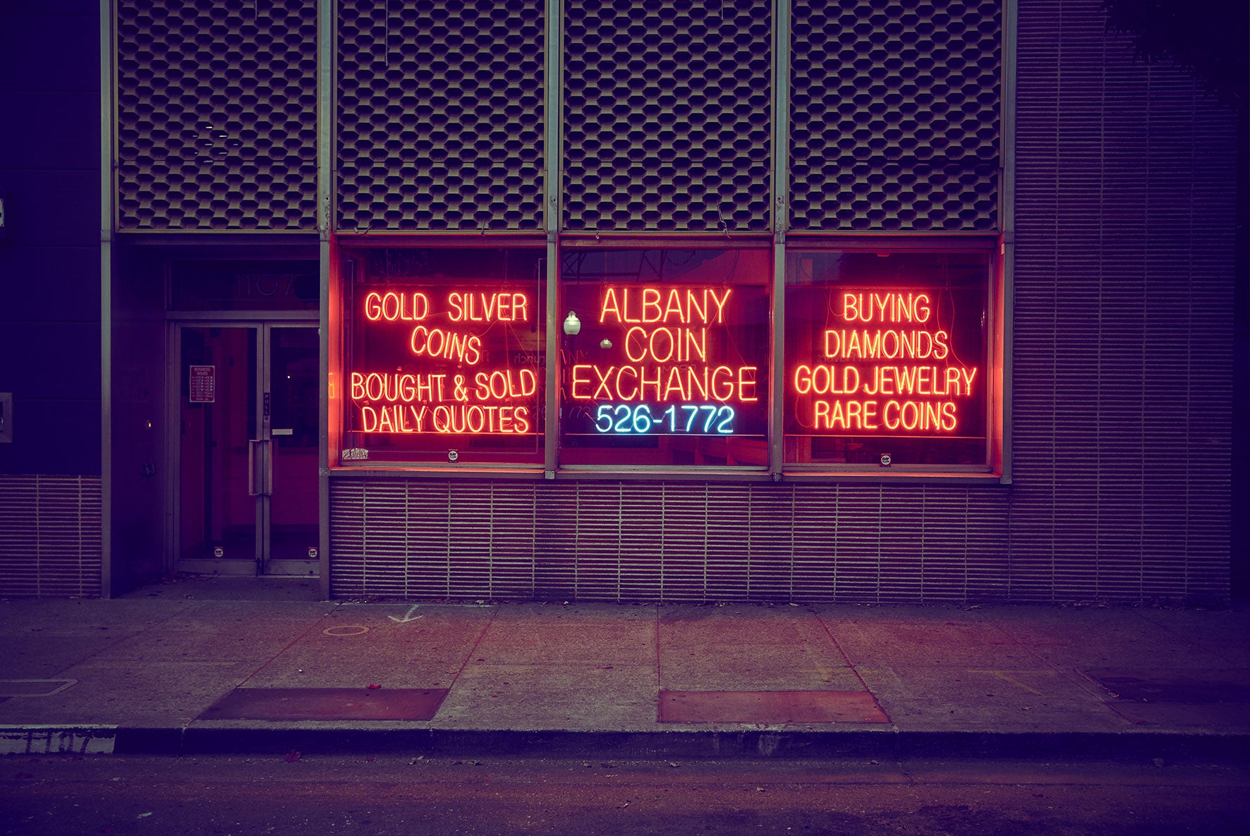 Dusk street view of “Albany Coin Exchange” storefront with red neon lettering, blue phone number glow, honeycomb facade panels, and an empty sidewalk.Fine art photography by Frank Kayser – Thrift Shop, San Francisco 2018.