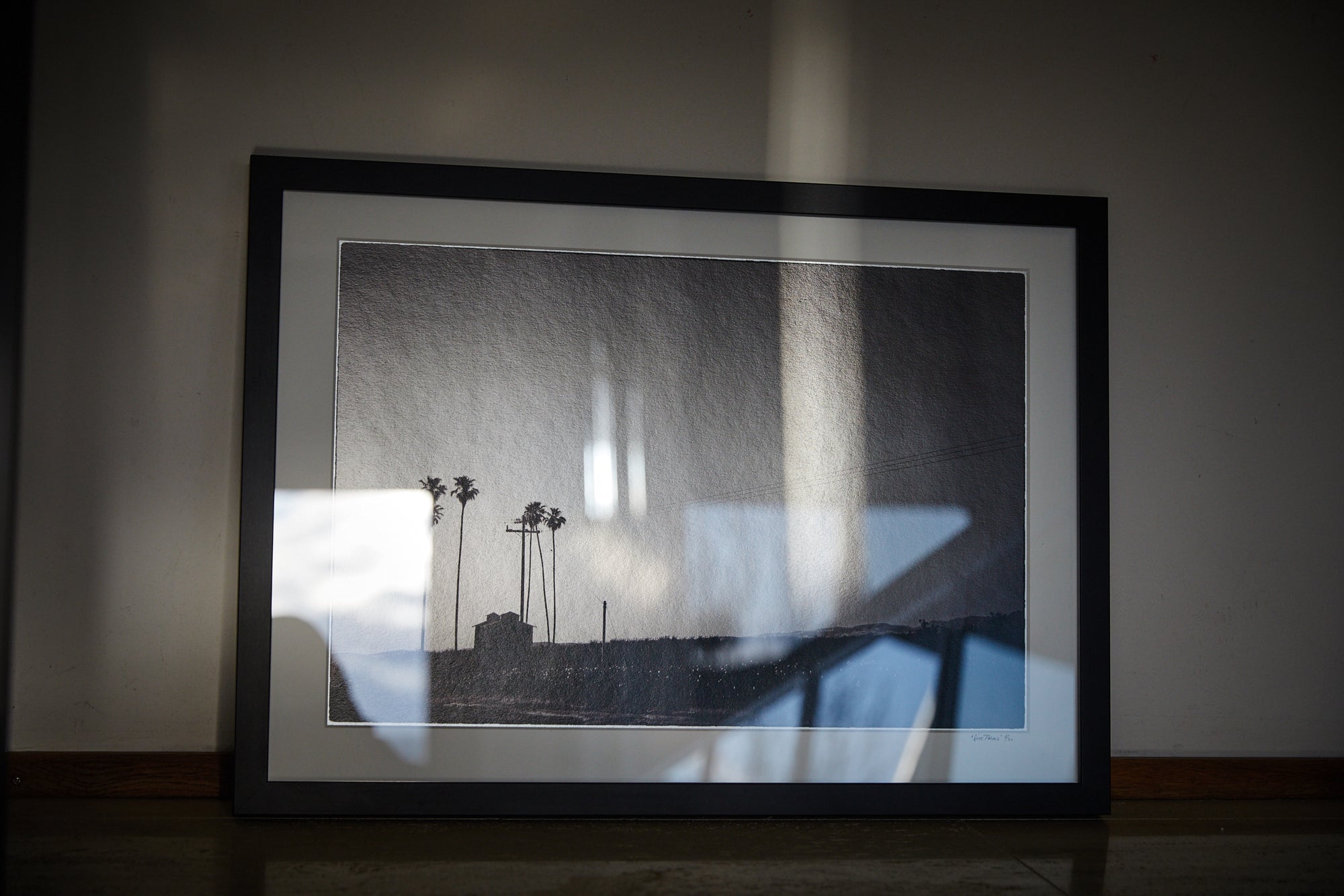 Framed black-and-white print of a minimalist landscape—slender palms, a small hut and a lone utility pole on the horizon—light reflections across the glass.