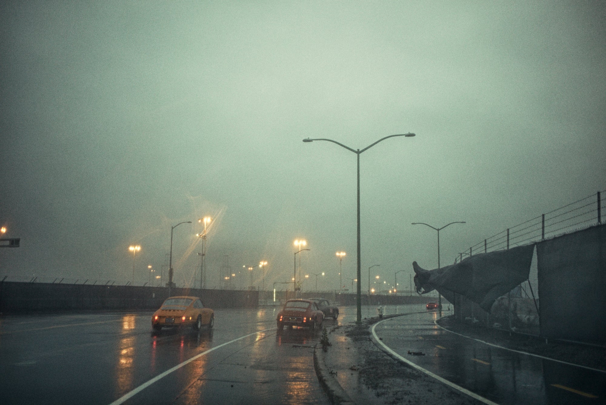 Rain-soaked industrial roadway at dusk with two classic Porsche 911s, sodium streetlights, and a torn fence tarp flapping in wind.