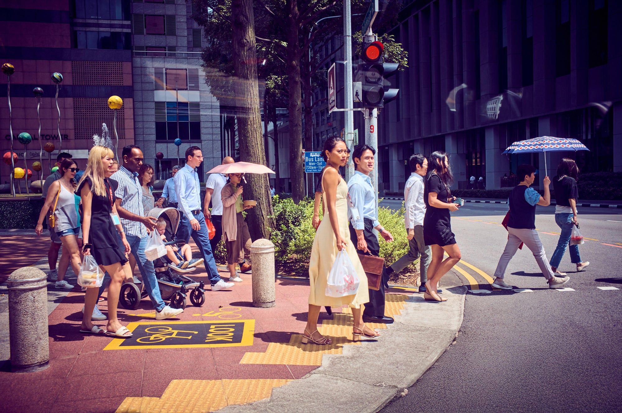 Diverse pedestrians at a Singapore crosswalk in midday sun—umbrella, stroller, and office towers in view.