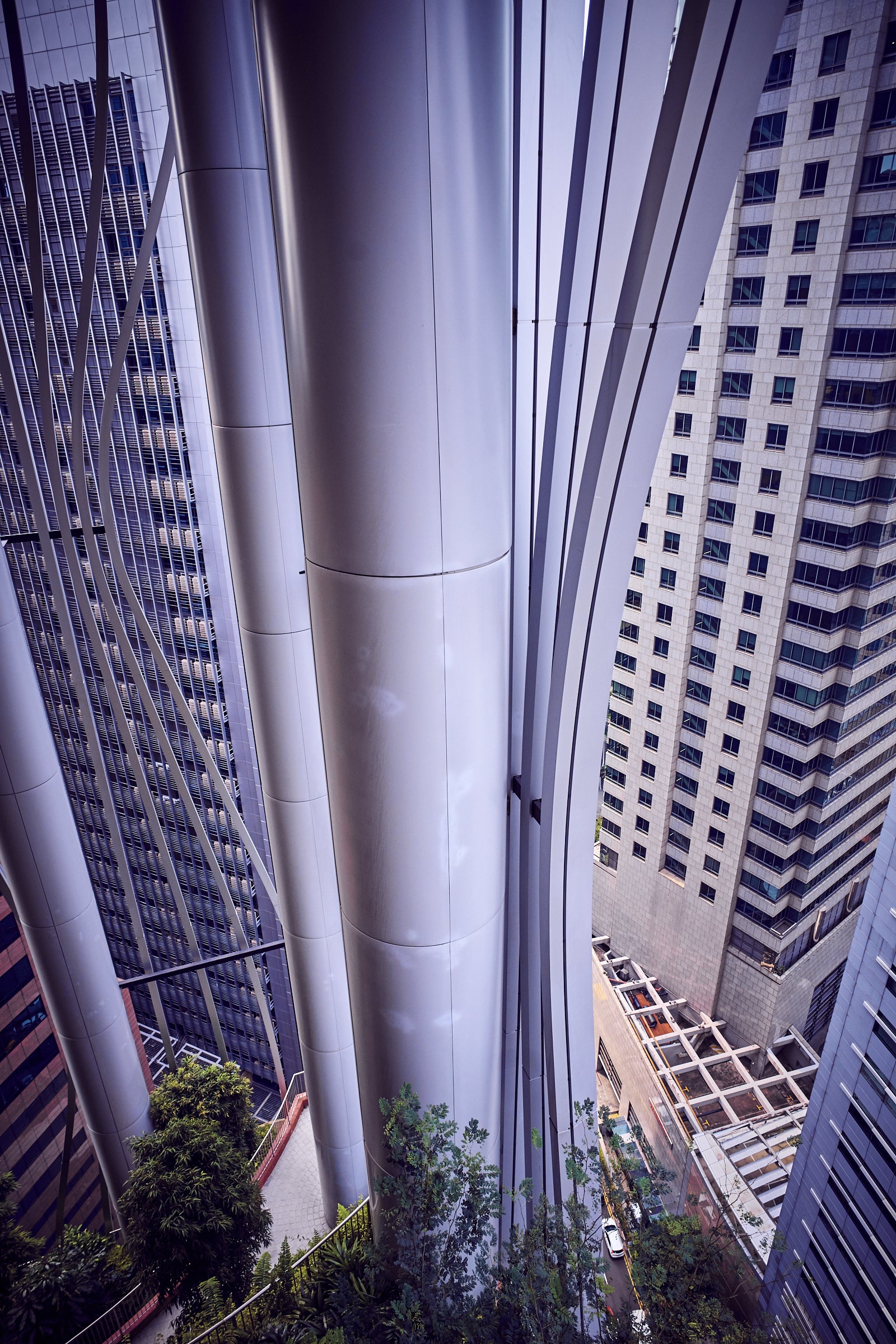 Curved silver structural columns with vertical fins between high-rise facades, greenery below, Singapore.