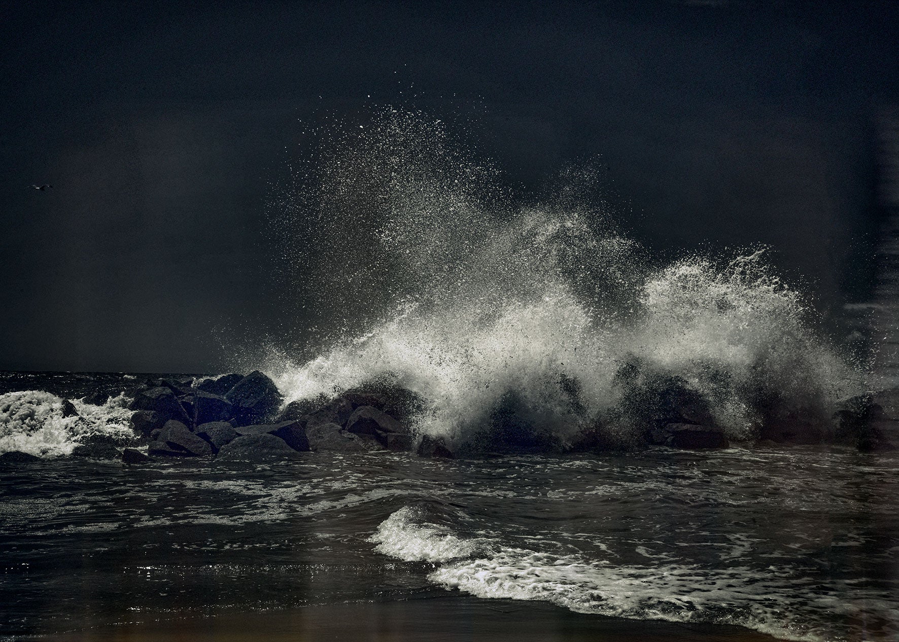Powerful ocean wave exploding over dark rocks, salt spray frozen against a deep horizon.